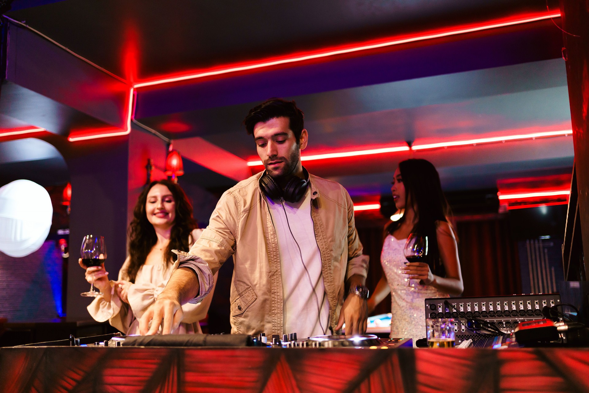 A DJ performs in a vibrant nightclub, mixing tracks while two stylish women enjoy drinks. Neon lights, energetic atmosphere, and music set the mood for a lively nightlife experience.
